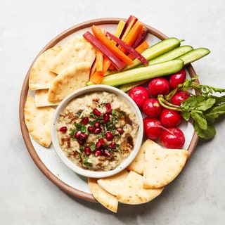 Pured eggplant in a bowl topped with pomegranate seeds and surrounded by cucumbers radishes carrots and pita chips.