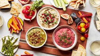 Three bowls of mutabal surrounded by figs tomatoes cucumbers asparagus carrots pita and pomegranates.